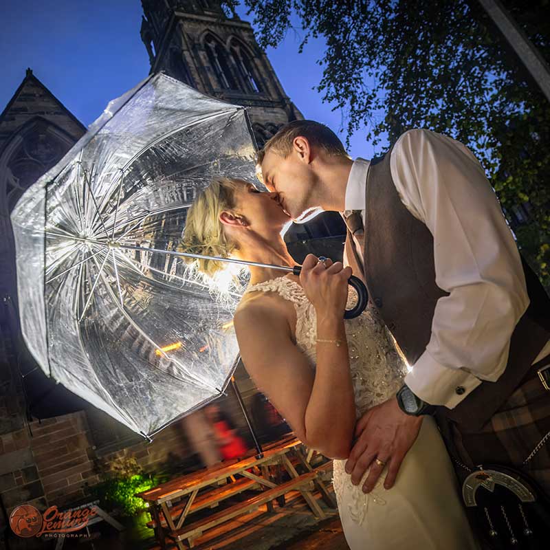 Romantic evening wedding portrait of a couple kissing beneath an umbrella outside the illuminated Cottiers venue in Glasgow.